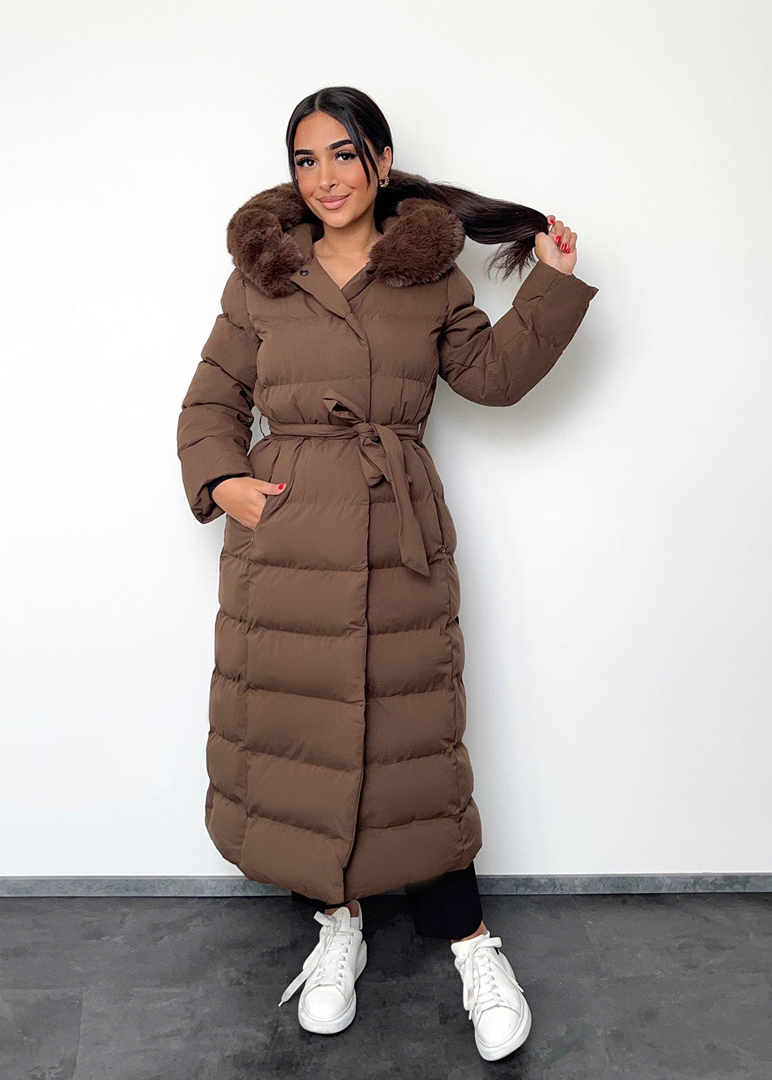 Modell trägt einen langen Damen Steppmantel mit Kapuze und Taillengürtel – NAHA Wintermode