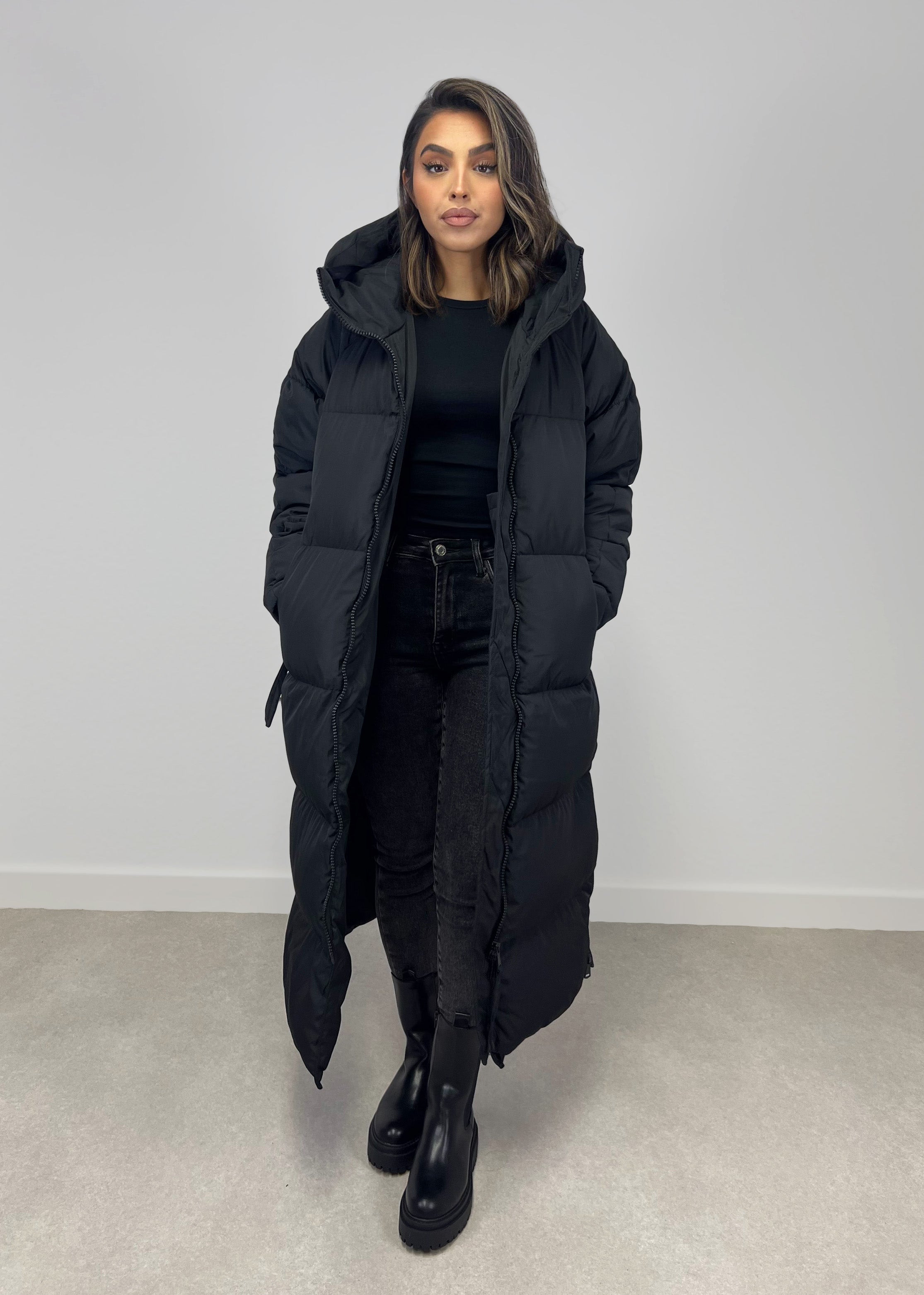 Modell trägt einen langen Damen Steppmantel mit Kapuze und durchgehendem Reißverschluss – NAHA Fashion