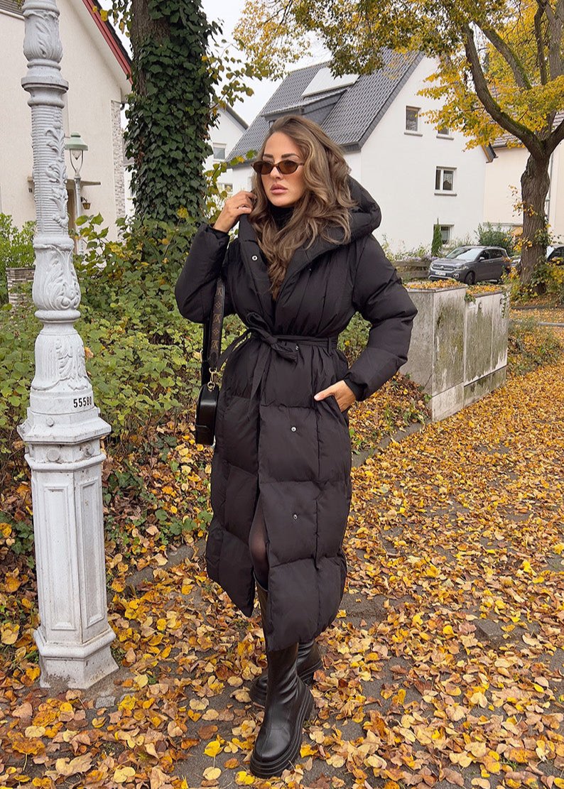 Modell trägt langen schwarzen Damen Steppmantel mit Kapuze und Taillengürtel, kombiniert mit Stiefeln und Sonnenbrille im herbstlichen Streetstyle – NAHA Winterjacke